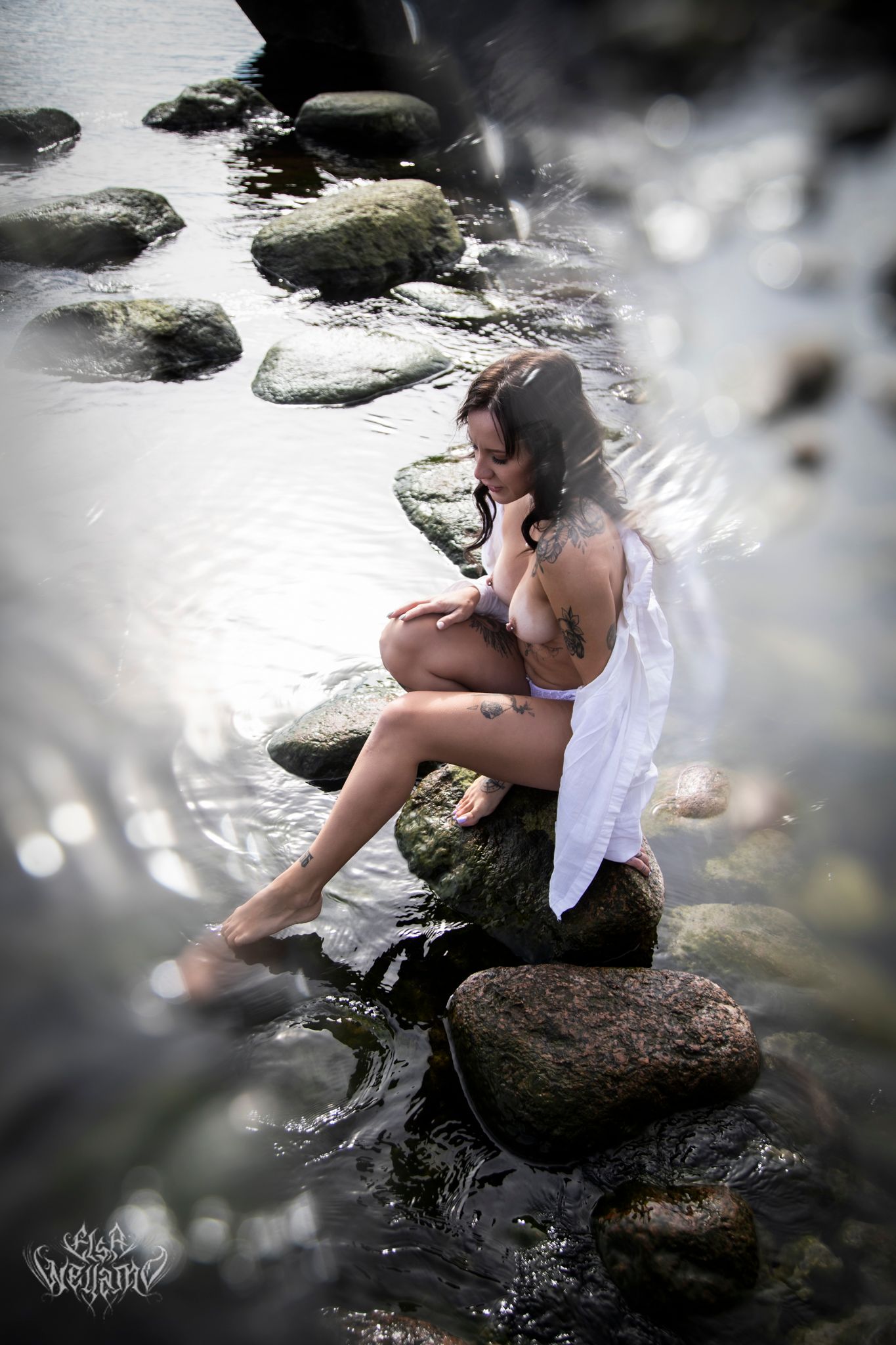 Artistic portrait of someone in white sitting on rocks by the water's edge with dreamy bokeh effects in nature.