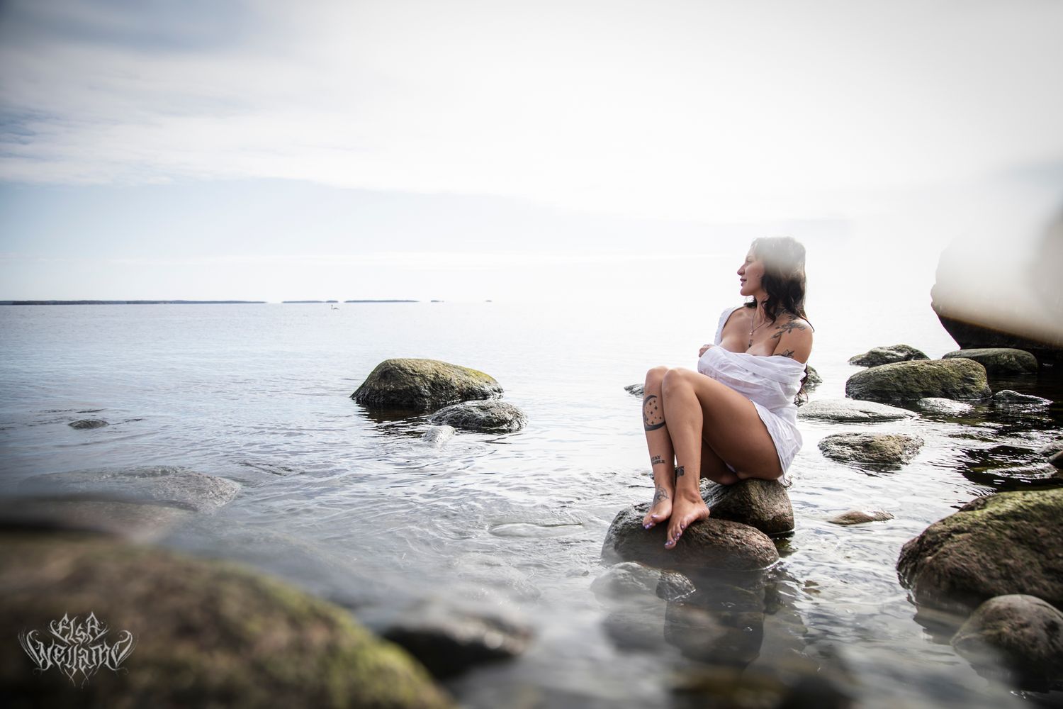 A lone figure sits on coastal rocks at the water's edge during a tranquil seascape scene.