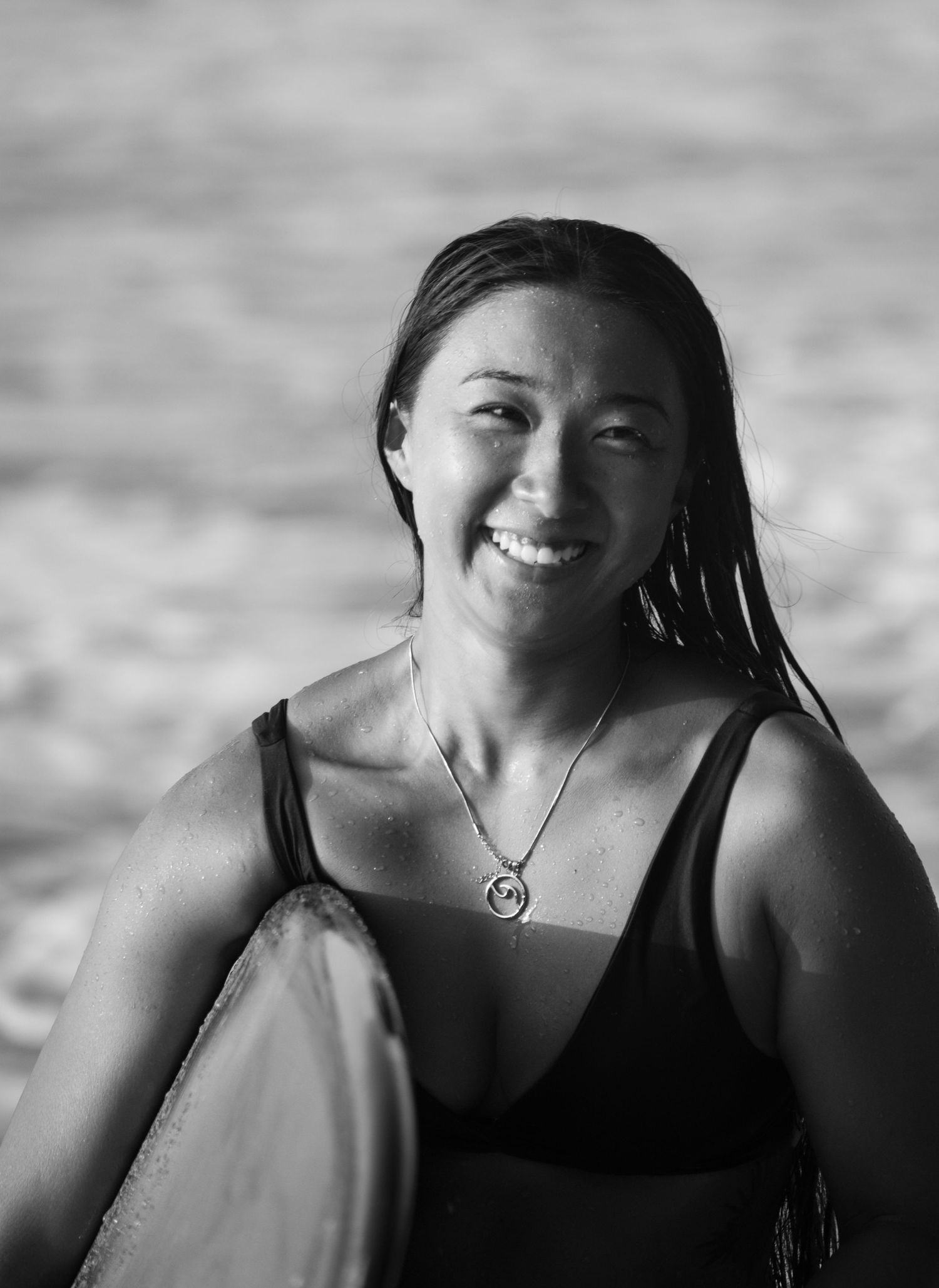 Black and white portrait photo of a happy surfer in a tank top with wet hair and necklace against a blurred background.