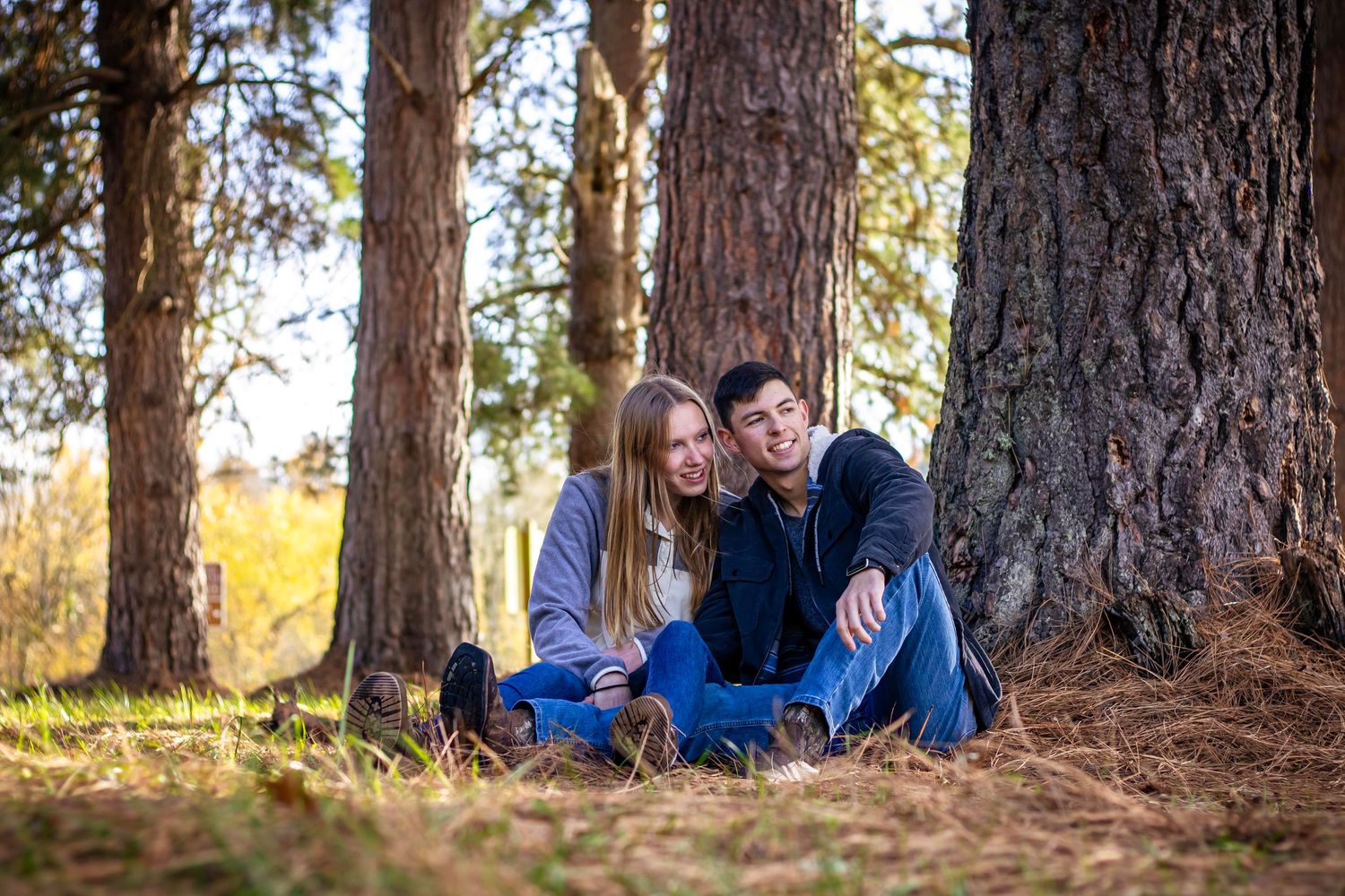 man and woman sit together under trees looking to the right as light seeps through trees behind them