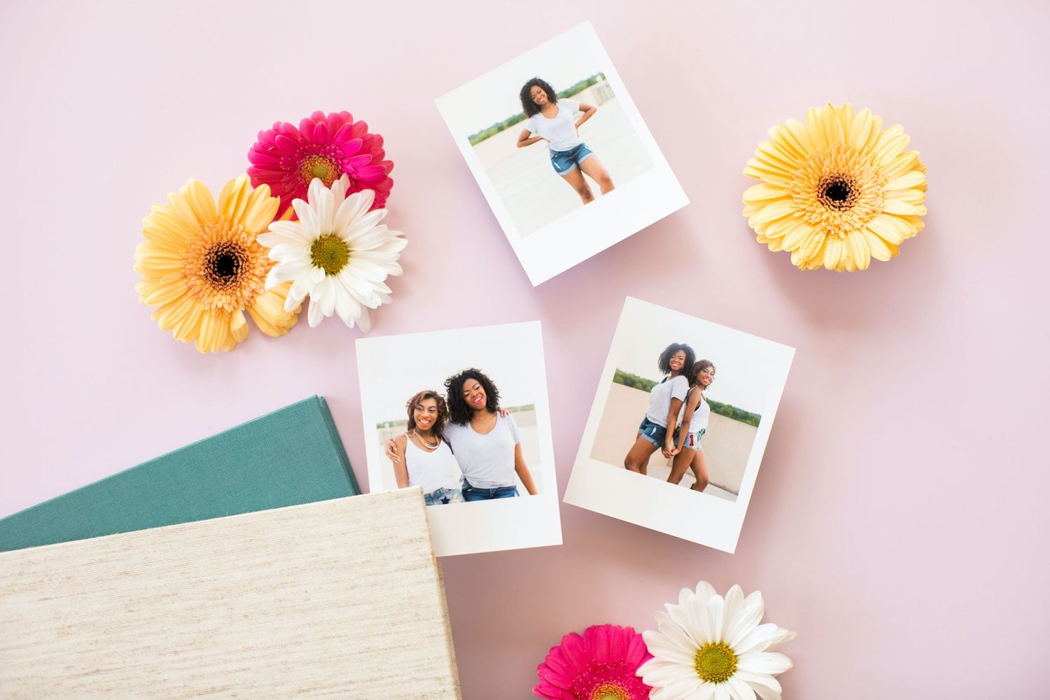 Colorful gerbera daisies and polaroid photos arranged with teal and cream notebooks on pink background.