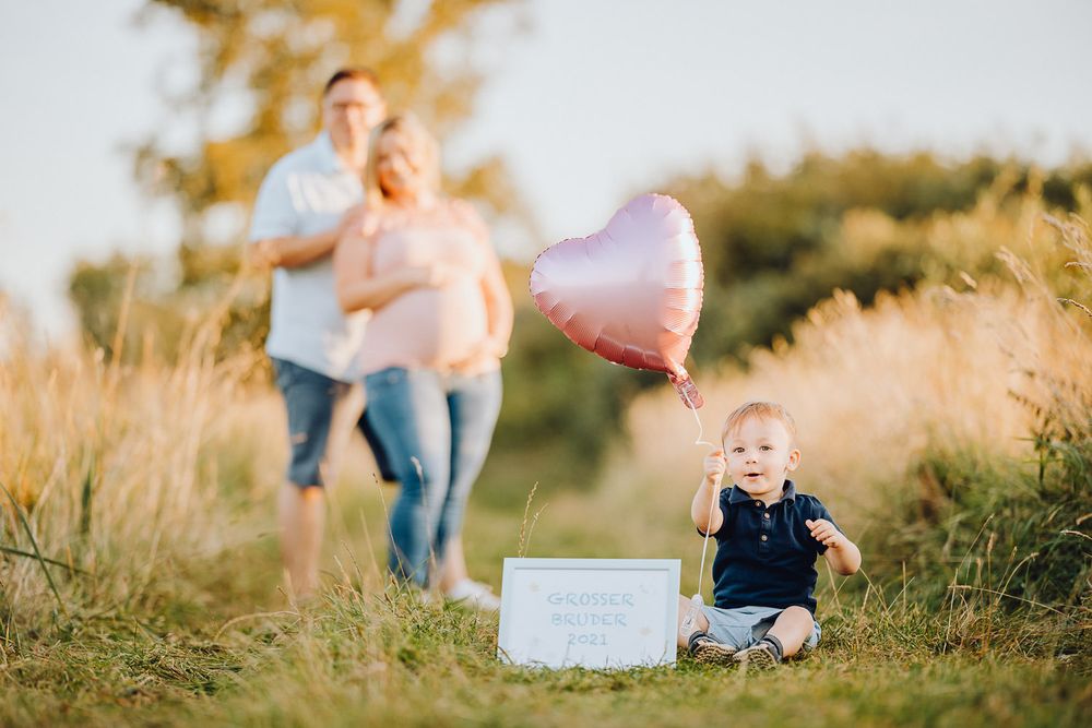 Gender reveal setting with toddler sitting by balloon and box in grassy field.