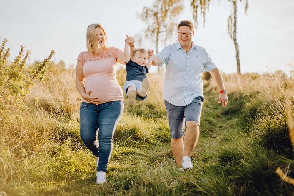 Family playing and swinging toddler between them while walking on grassy path.