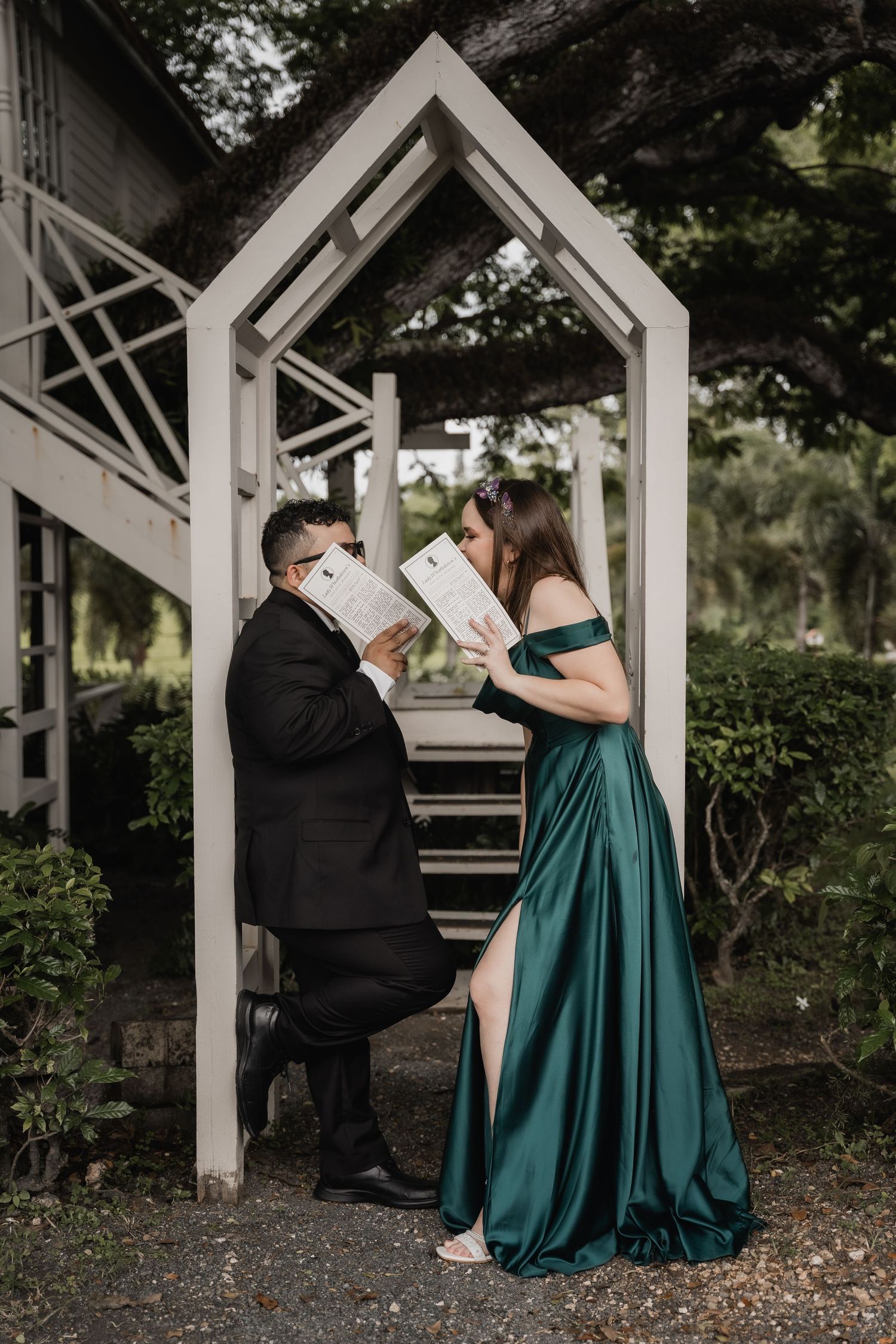 A couple in formal attire share a romantic moment under a white gazebo, with her wearing an elegant emerald green gown.