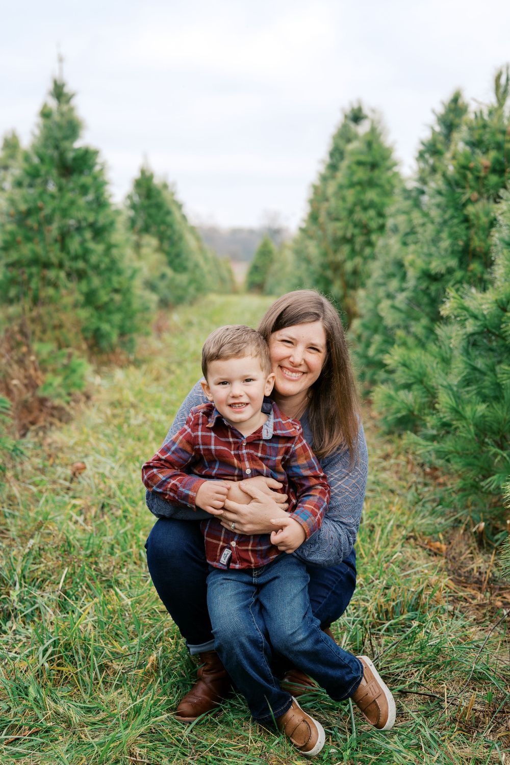 A parent and child share a warm embrace while kneeling among Christmas trees.