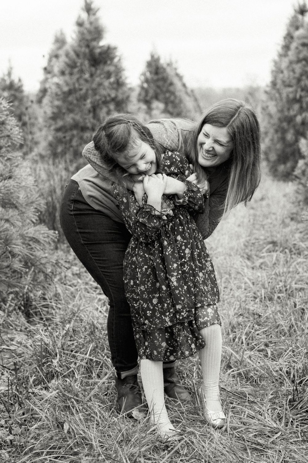 Two people share playful moments together in a field with evergreen trees in black and white.