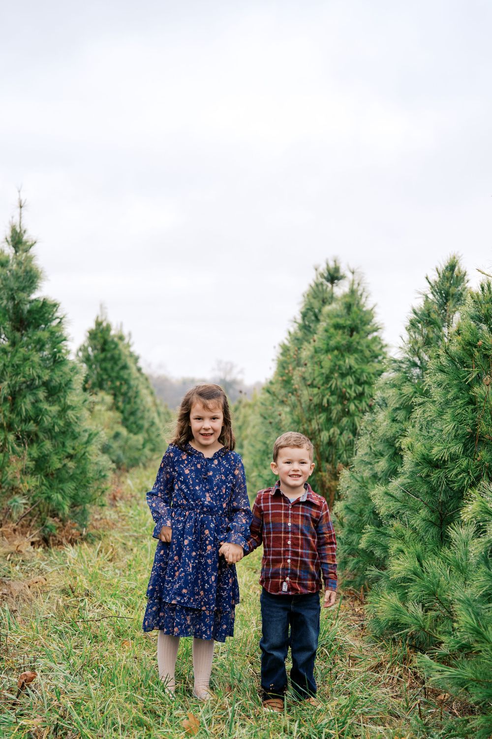 Two children stand together smiling among evergreen trees at a Christmas tree farm.