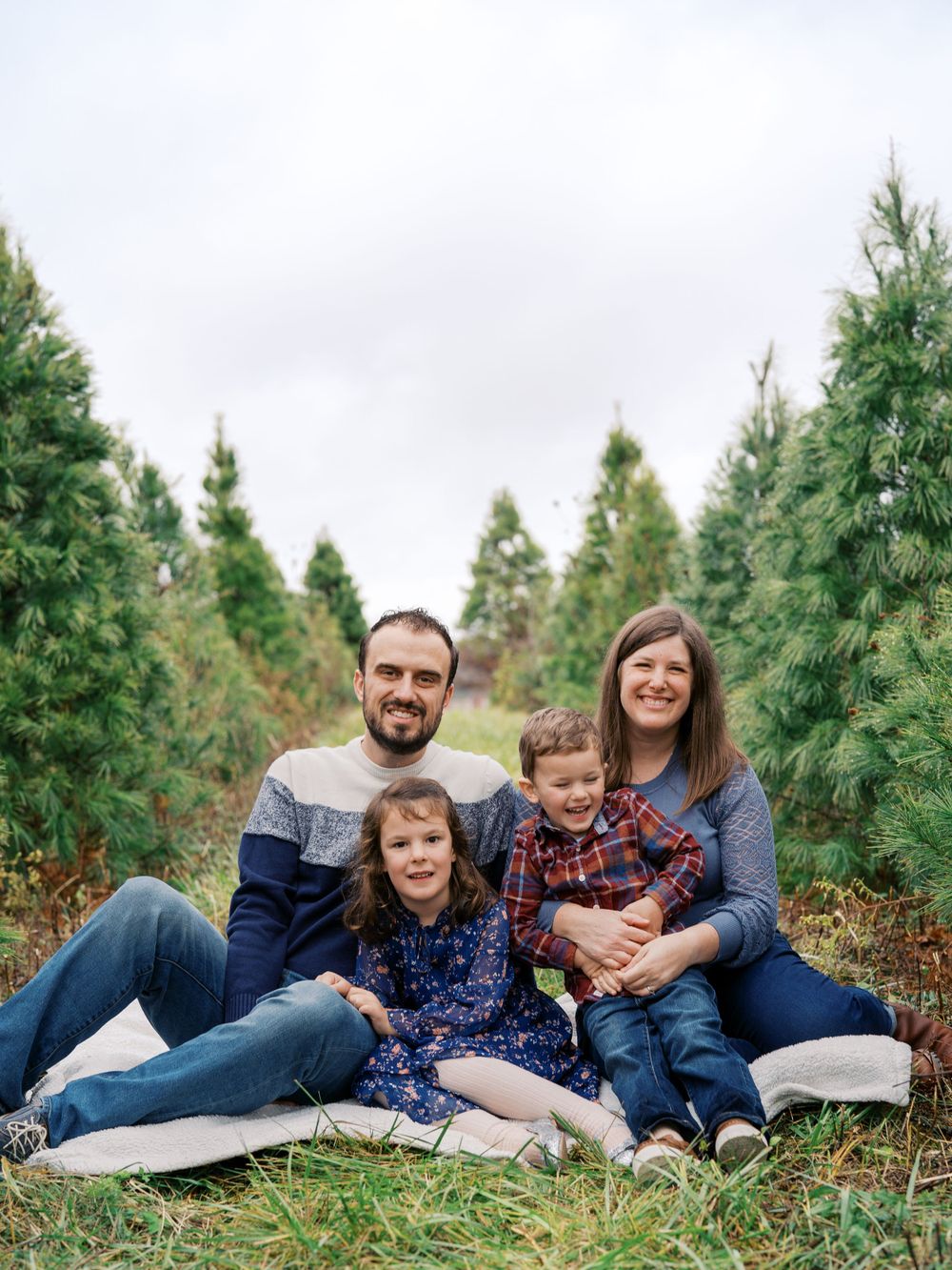 Family relaxes together on ground among evergreen trees during outdoor photo session.