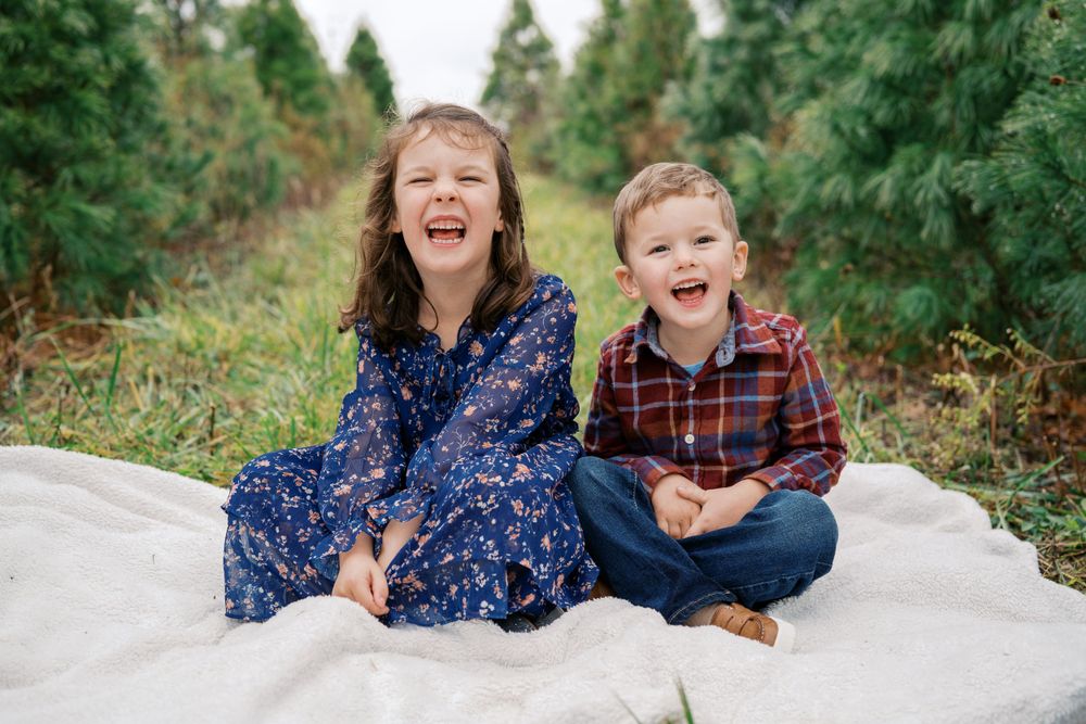 Playful moments captured of children sitting on white blanket among evergreen trees.