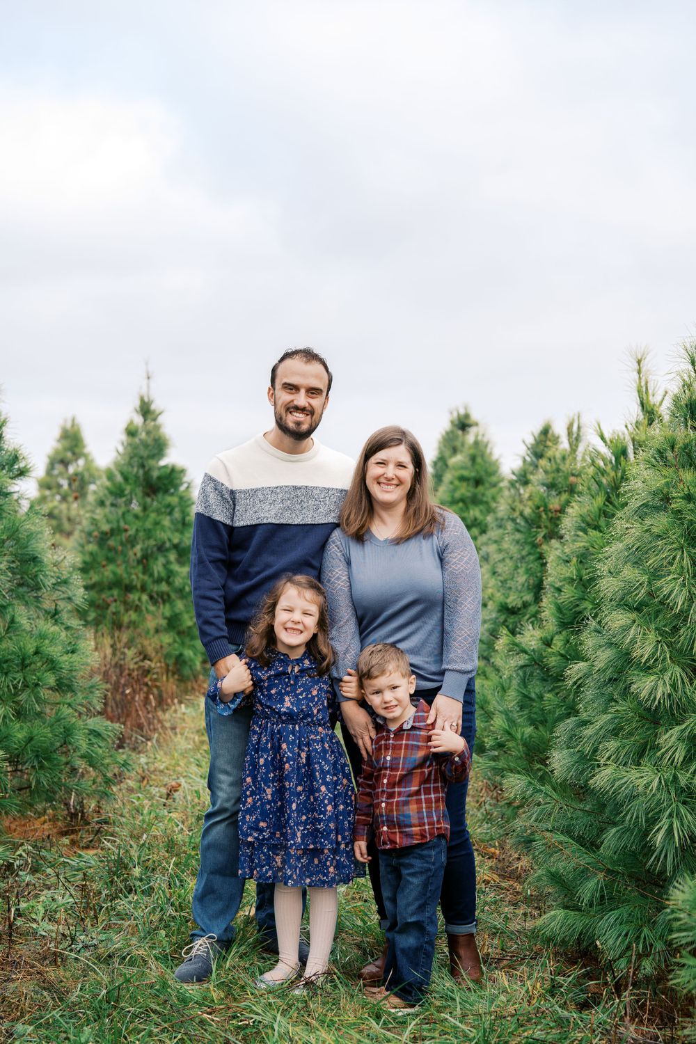 A family portrait with two children standing between rows of pine trees on a cloudy day.
