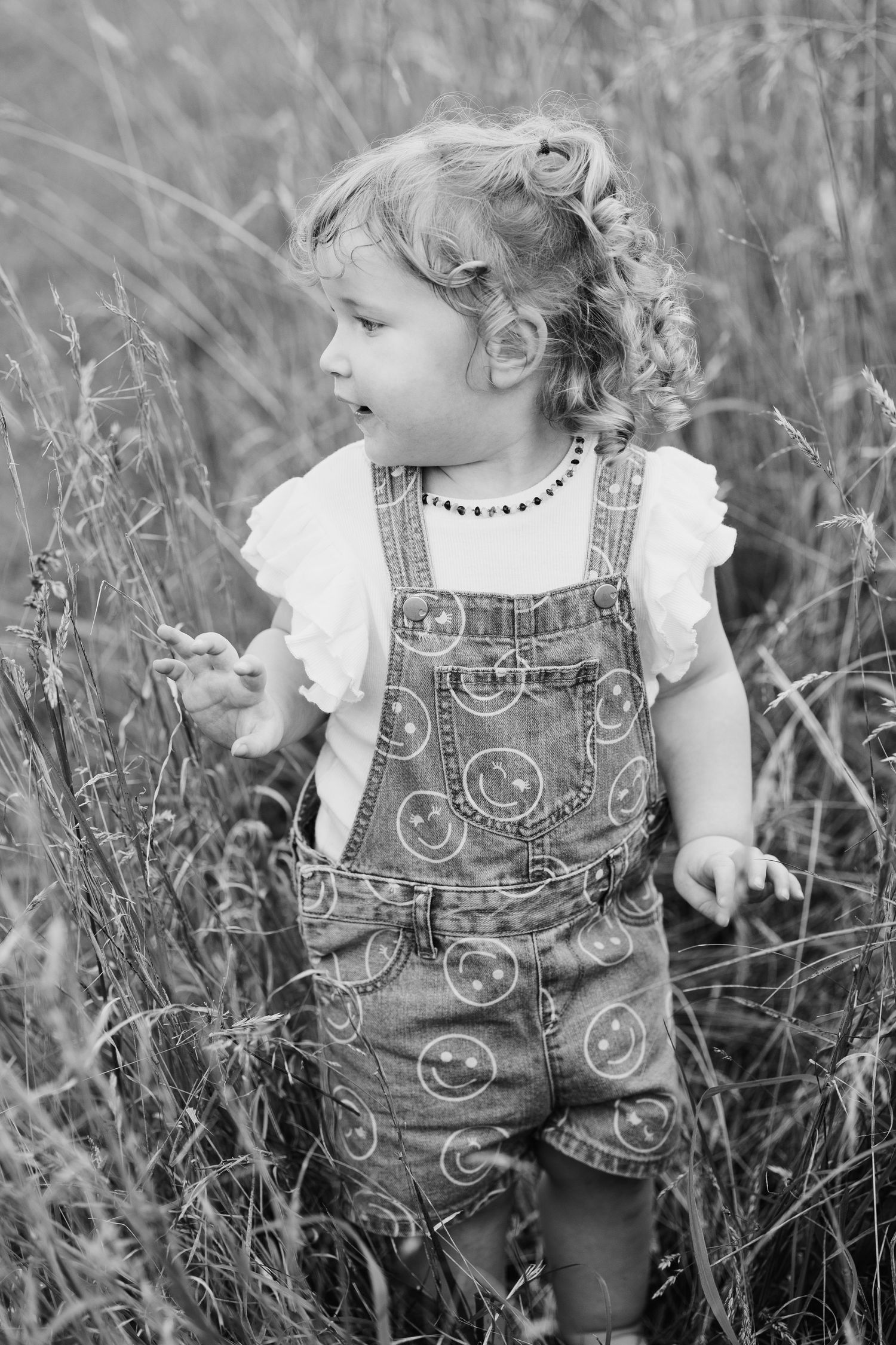 Young child in overalls and ruffled shirt exploring tall grass in black and white.