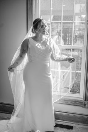 A bride in a flowing white dress poses gracefully by a sunlit window.
