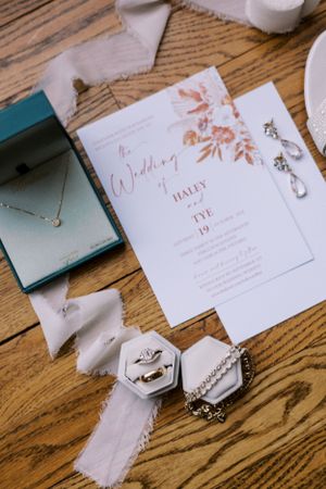 Wedding invitation with floral design alongside jewelry and accessories on wooden surface.