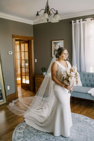 A bride holds a white bouquet in an elegant room with hardwood floors and gray walls.