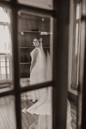 A bride in a sleek white dress poses through vintage window panes in black and white.