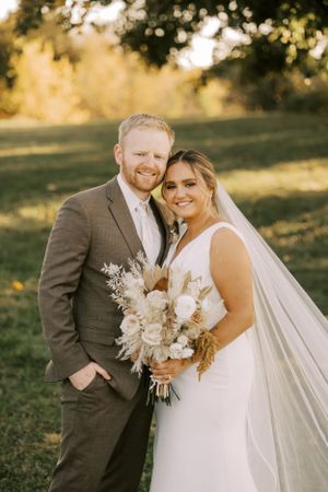 Newlyweds pose together in sunlit outdoor setting with bride holding neutral-toned floral bouquet.