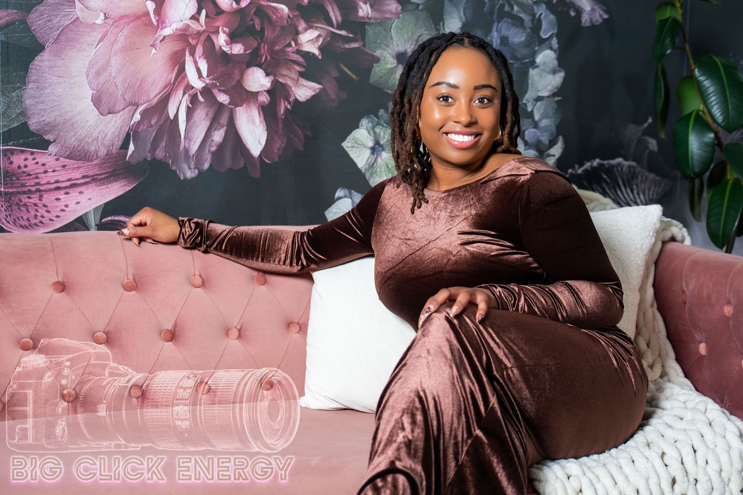 With a radiant smile, a young Black woman lounges on a pink velvet couch, her chocolate gown draped elegantly, florals behind her.
