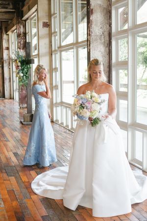 Bride in white strapless gown holds pastel bouquet while bridesmaid in blue gown looks on in elegant venue.