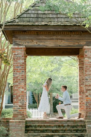 Man proposes on one knee to woman in white dress under rustic brick gazebo surrounded by greenery.