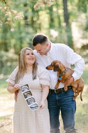 A smiling couple holds ultrasound photos and a dachshund dog in a sunlit forest for a pregnancy announcement. Engagement session at Rowan