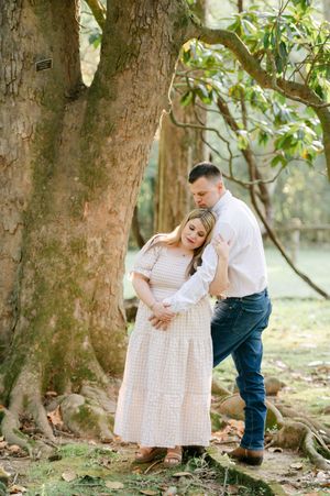 A couple embracing tenderly under a large tree in a lush outdoor setting. Maternity session at Rowan Oak in Oxford, Mississippi.