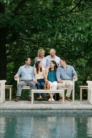 Extended family of six smiles together on white benches beside a pool surrounded by lush trees.
