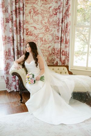 Bride in white gown with veil sits gracefully on vintage chaise lounge before red toile curtains.