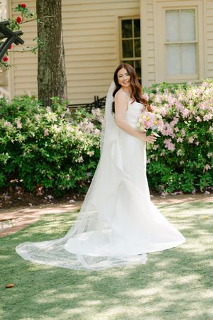 Smiling bride in white gown with lace veil poses before blooming pink azalea bushes outdoors.