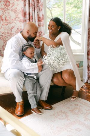 Happy family of three in white attire share tender moment with baby's breath bouquet indoors.