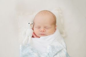 Peaceful newborn baby sleeps swaddled in a blue blanket with a small white teddy bear beside them.