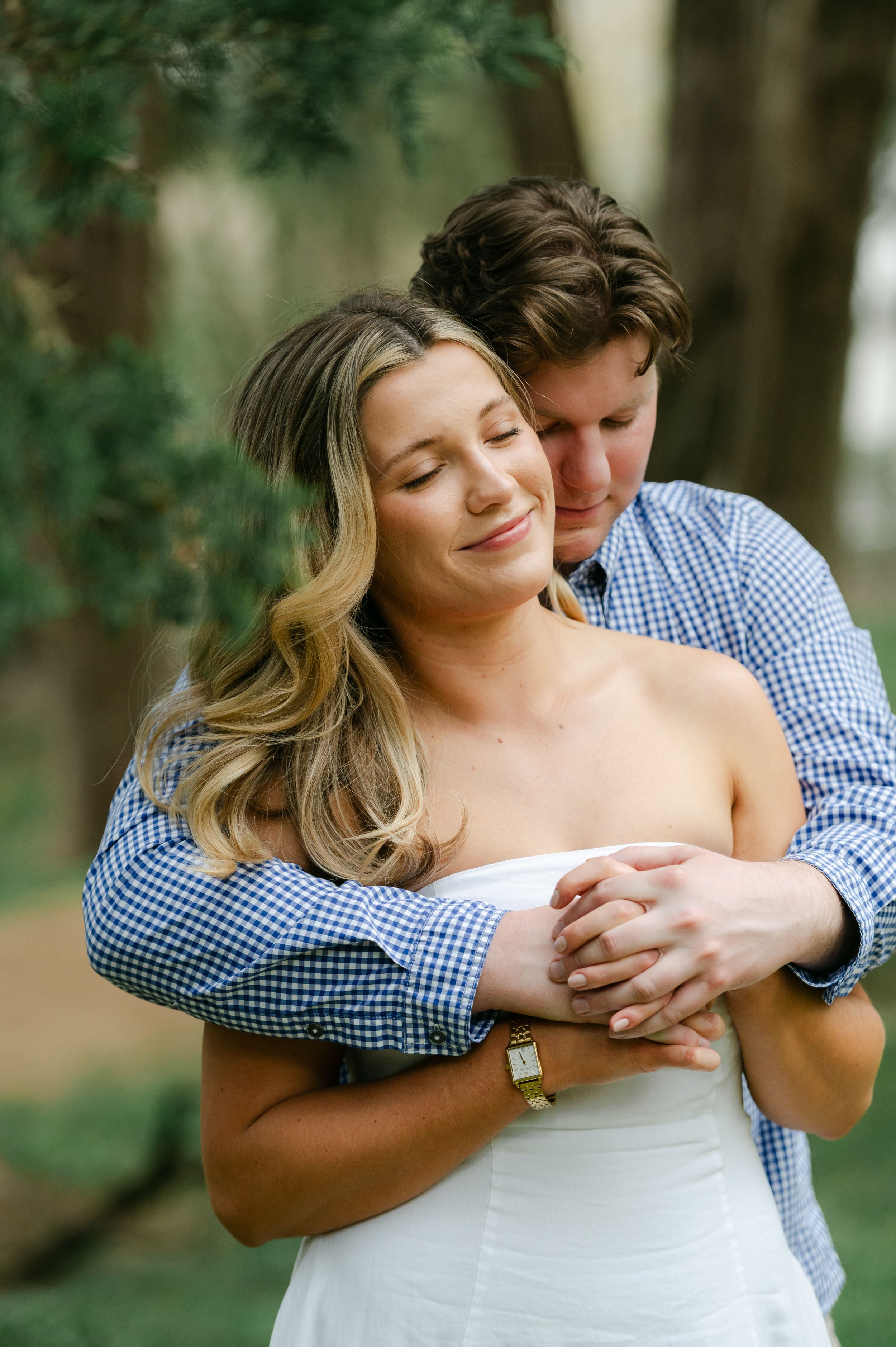 A man hugging a woman from behind as she smiles with eyes closed outdoors—engagement session at Rowan Oak in Oxford, Mississippi.