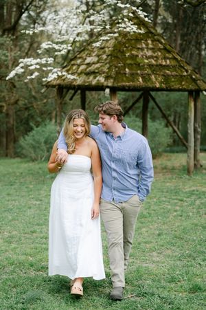 A smiling couple walking together on green grass near a moss-covered gazebo. Engagement session at Rowan Oak in Oxford, Mississippi.