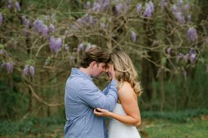 Couple touching foreheads romantically under blooming purple wisteria trees—engagement session at Rowan Oak in Oxford, Mississippi.