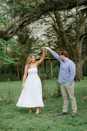 A man spinning a woman in a white dress under flowering dogwood trees outdoors. Engagement session at Rowan Oak in Oxford, Mississippi.
