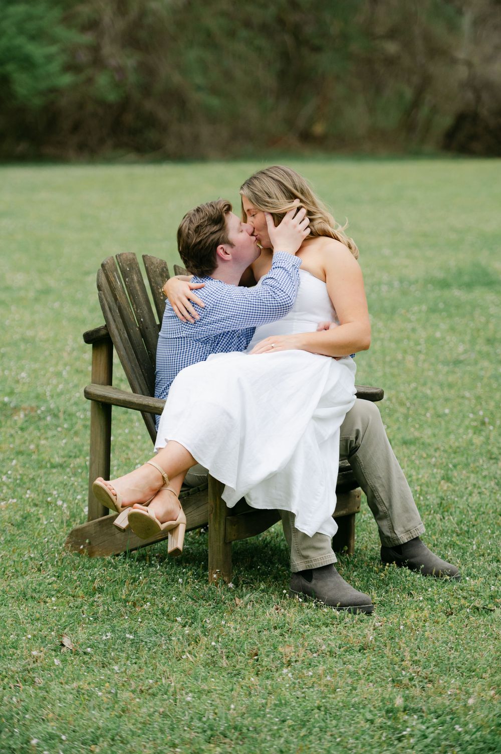 A couple sharing a kiss while sitting together in a wooden Adirondack chair. Engagement session at Rowan Oak in Oxford, Mississippi.