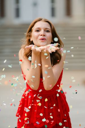 A woman in a red dress blows colorful confetti from her hands outdoors on thesteps. Ole Miss graduate celebrating graduation with a portrait
