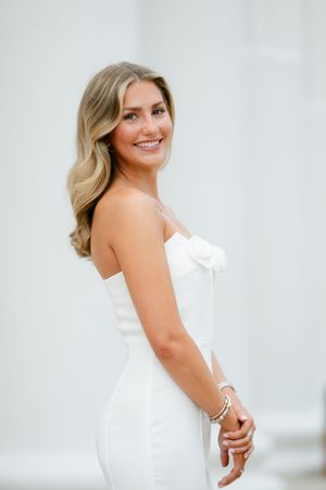 Smiling blonde bride in a strapless white gown with floral detail poses against a soft white background. Graduation portraits.