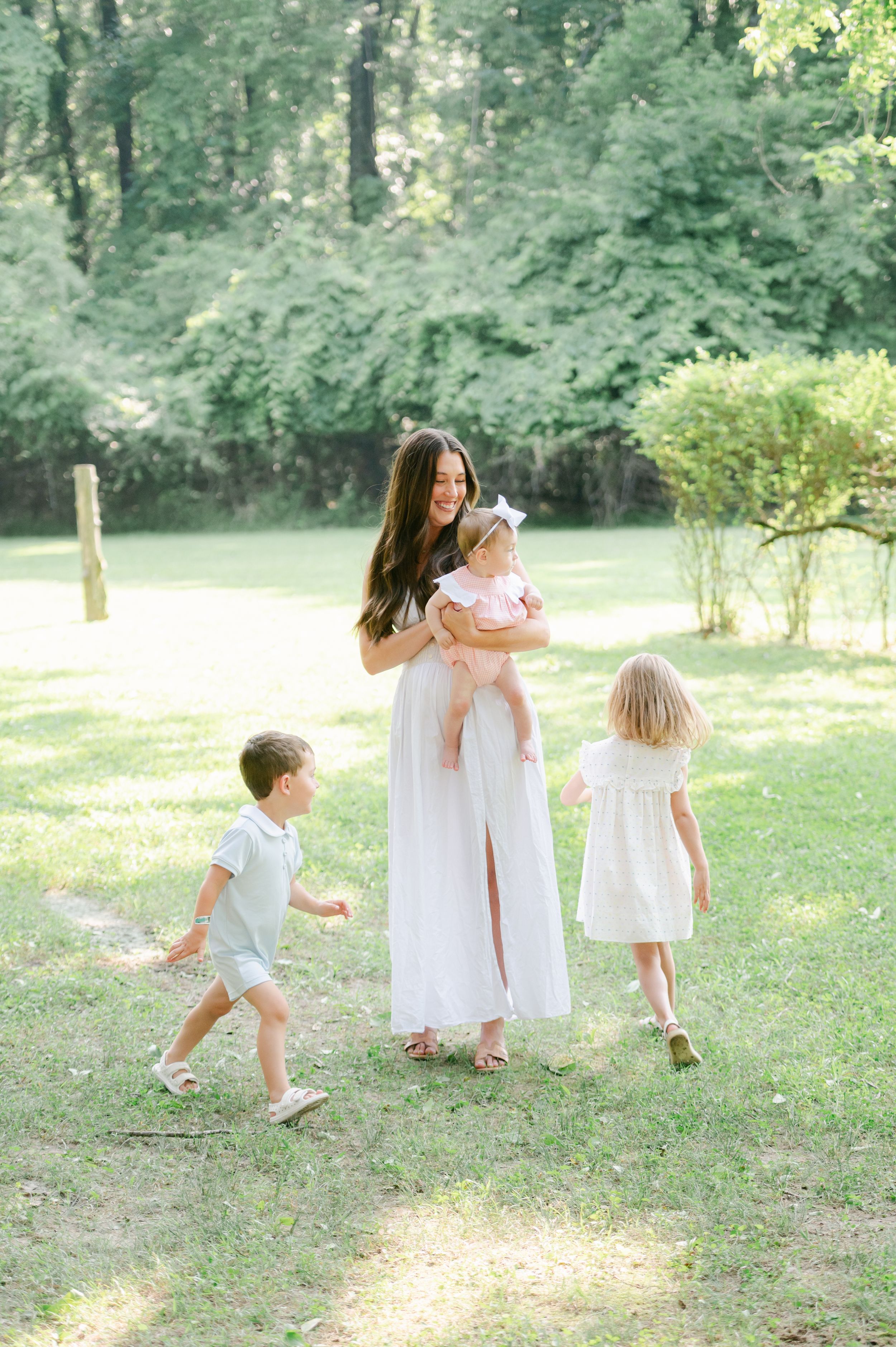 Brunette mother in a white dress holds her baby while two young children walk beside her outdoors.