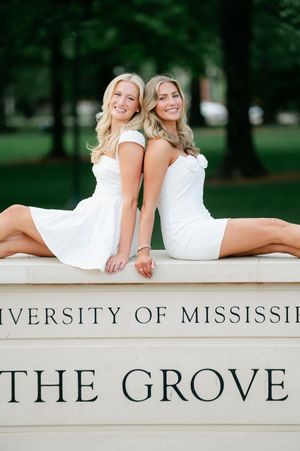Two smiling women in white dresses pose at the University of Mississippi The Grove sign for graduation.