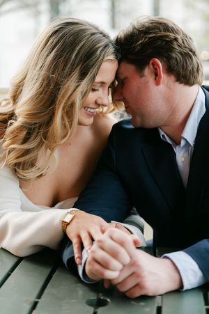 A smiling couple leaning close together, holding hands at an outdoor table. Engagement session in Oxford, Mississippi.