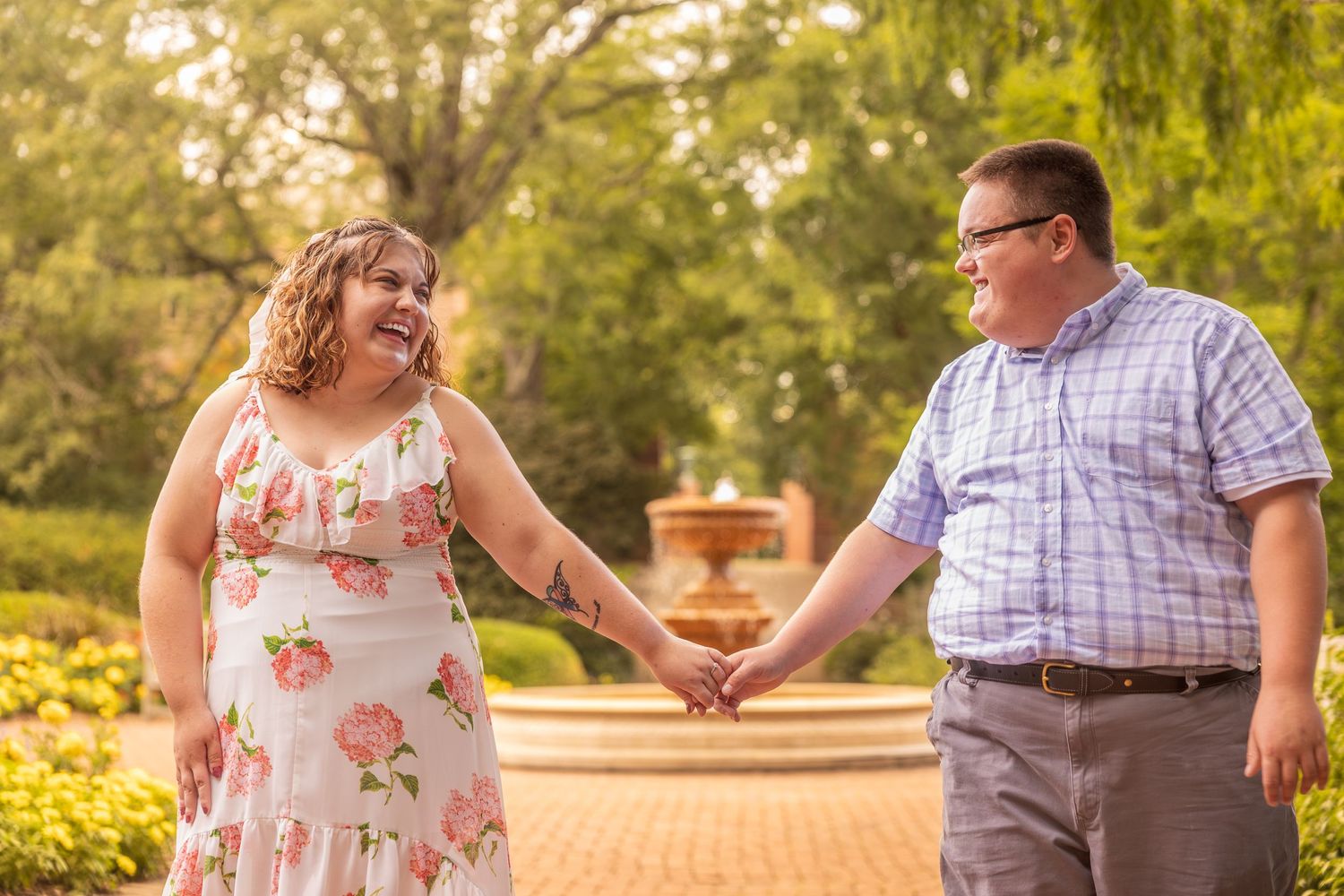 A couple holds hands in a romantic garden setting during sunset with trees and flowers in the background.