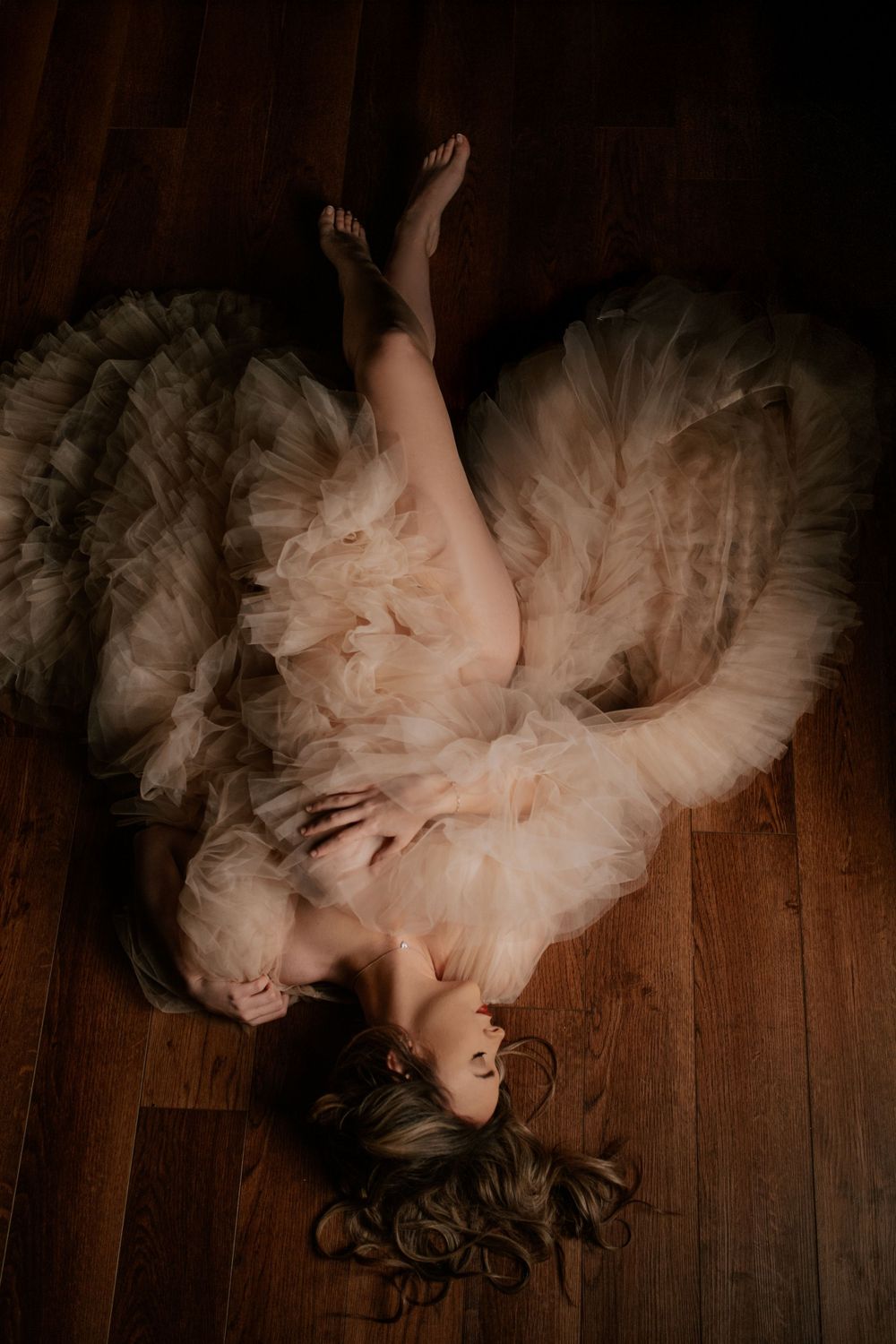 A dreamy portrait of someone lying on a wooden floor wearing a flowing cream-colored tulle dress.