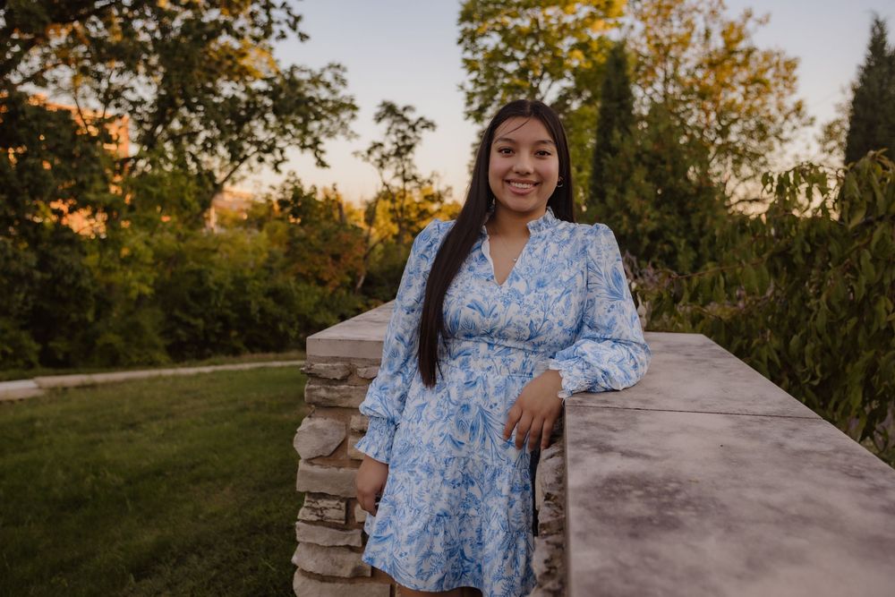 A person in a blue floral dress poses outdoors at sunset against rustic stone walls and autumn foliage.