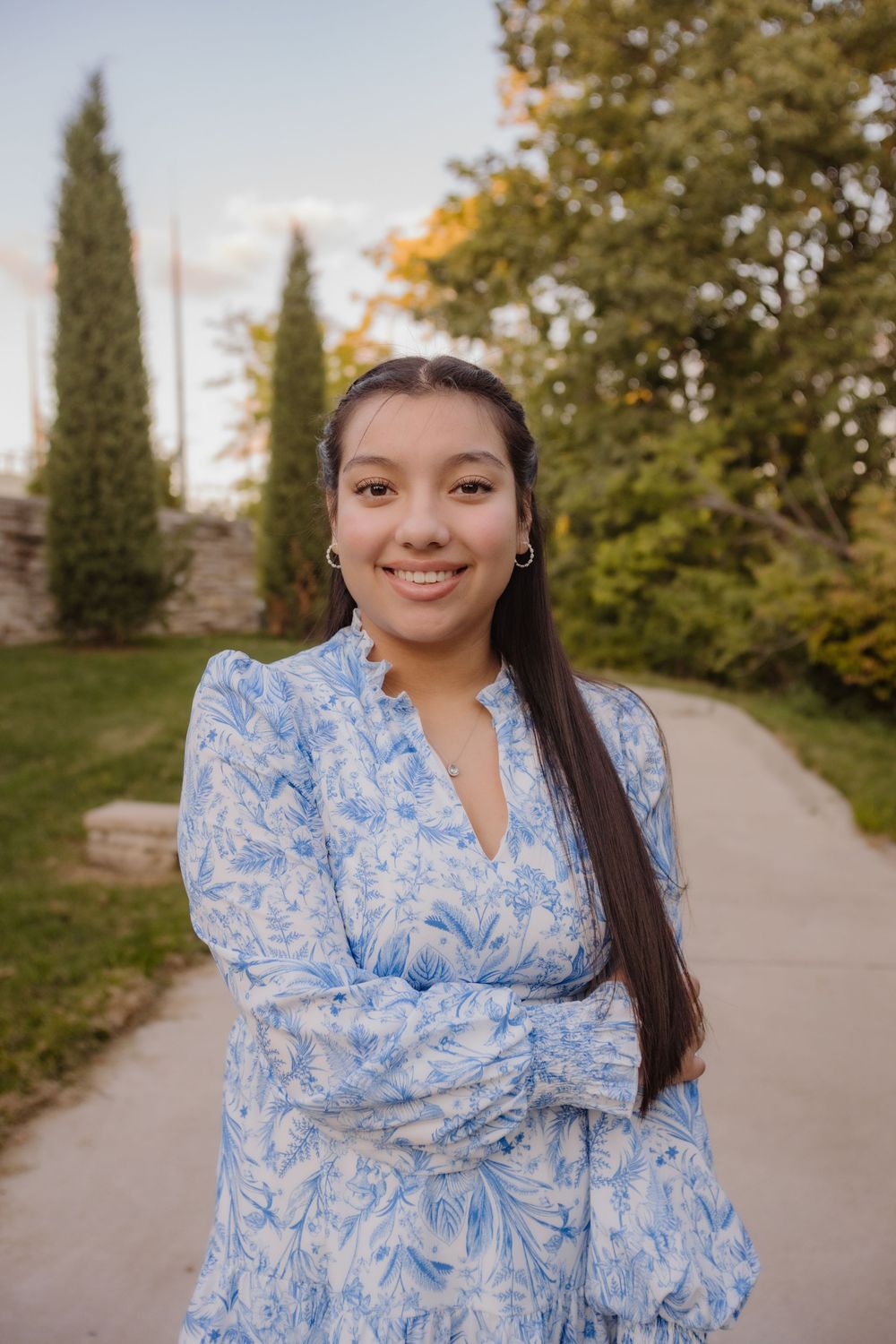 Someone wearing a blue and white floral dress poses on a garden path with cypress trees and autumn foliage in background.