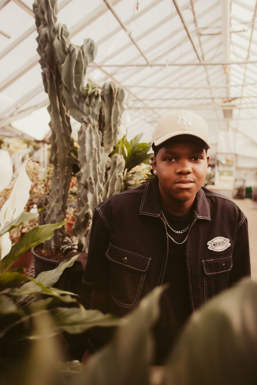 Person wearing a black jacket and baseball cap poses in a sunlit greenhouse surrounded by plants and hanging vines.