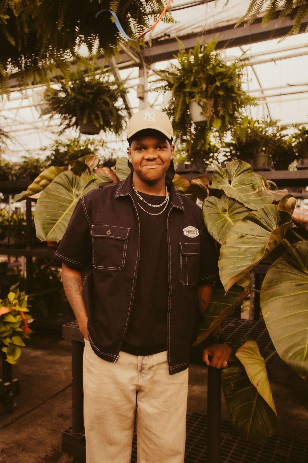 Someone wearing a dark jacket and light pants poses among lush tropical plants in a greenhouse setting.