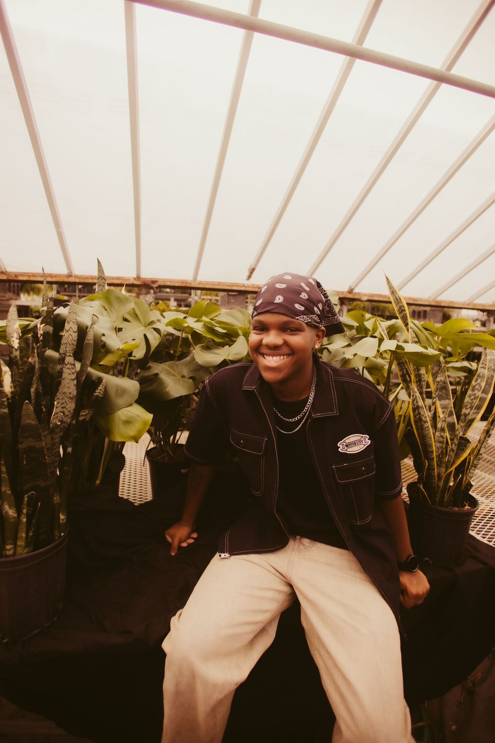 Person in casual attire poses among potted plants in a greenhouse with translucent roof panels.