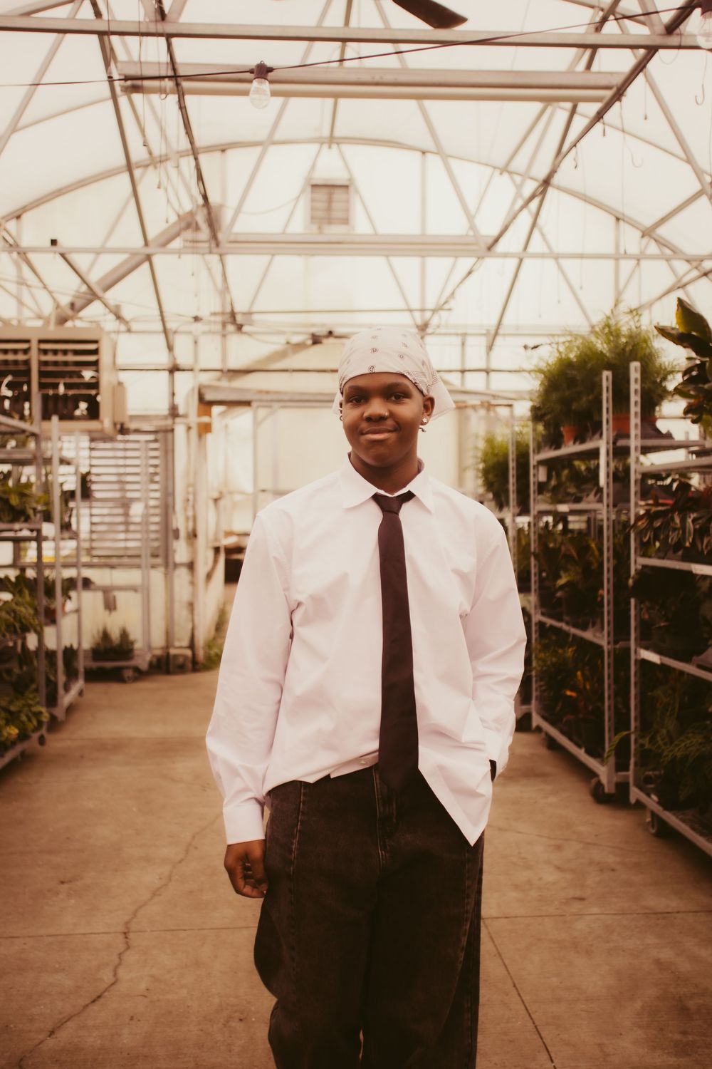 Person in formal white shirt and black tie stands in long greenhouse corridor with glass ceiling.