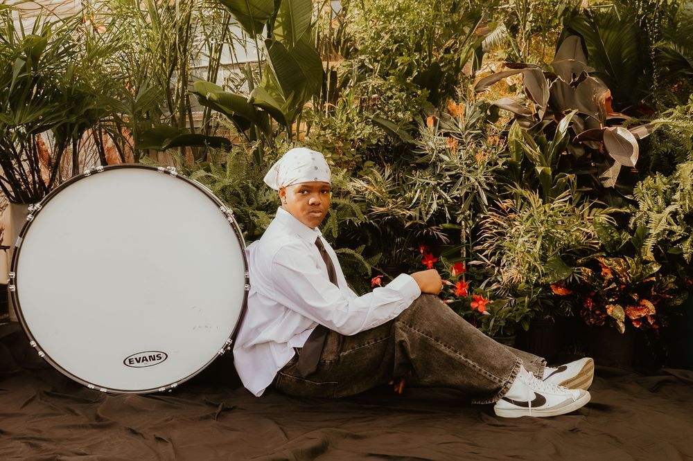 Someone in white rests beside a large white drum amid lush greenhouse plants and foliage.