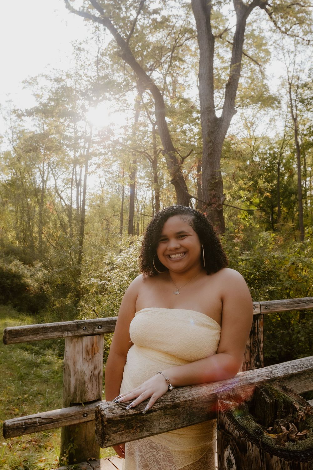 A person in a white dress poses against a rustic wooden fence surrounded by sunlit autumn trees in a forest setting.